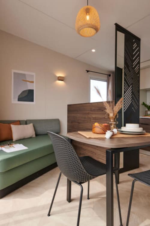 Intérieur contemporain d'un mobil-home 1 chambre au Le Maquis Plage avec canapé vert, bureau en bois, chaise noire et luminaire suspendu en rotin