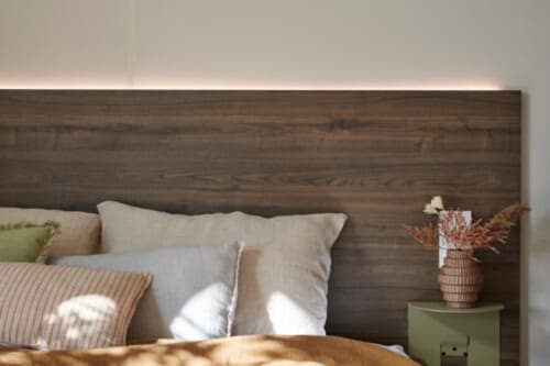 Chambre moderne au Maquis Plage avec tête de lit en bois foncé, oreillers crème, coussins rayés et fleurs en pot sur fond vert
