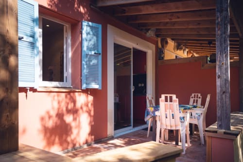 Terrasse ensoleillée de la villa Le Maquis Plage à Poggio-Mezzana avec table de repas, chaises blanches et façade aux murs rouge vif