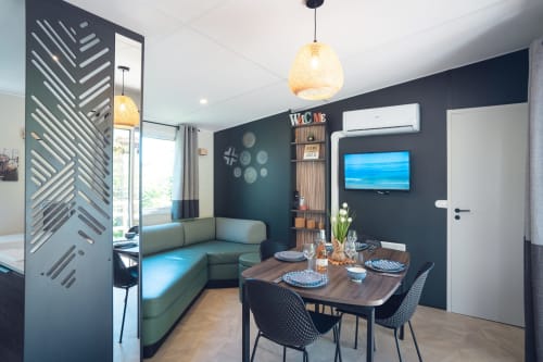 Espace de vie Mobil-Home Yuka moderne au Maquis Plage avec table à manger, canapé vert, télévision et luminaires en suspension tressés