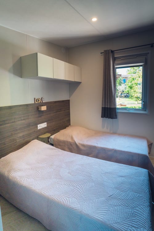 Chambre lumineuse avec deux lits jumeaux, tête de lit en bois foncé, placards blancs de rangement et fenêtre sur le jardin du Maquis
