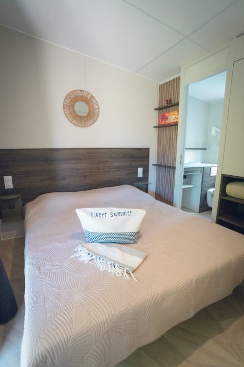 Chambre moderne au Maquis Plage avec lit queen-size, miroir tressé, tête de lit en bois et salle de bains attenante