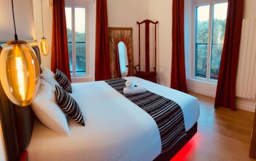 Chambre élégante du Moulin du Guy avec lit queen-size, chemin de lit rayé, luminaire suspendu, rideaux rouges et vue sur le lac