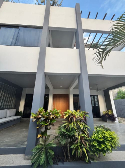 Entrada moderna de la Casa Tsuki en Puerto Escondido con muros blancos, pilares altos, puerta de madera y vegetación tropical