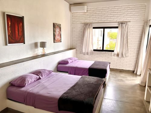 Moderna habitación en Casa Tsuki, Puerto Escondido, con ropa de cama morada, paredes de ladrillo blanco y ventanas con vista al mar