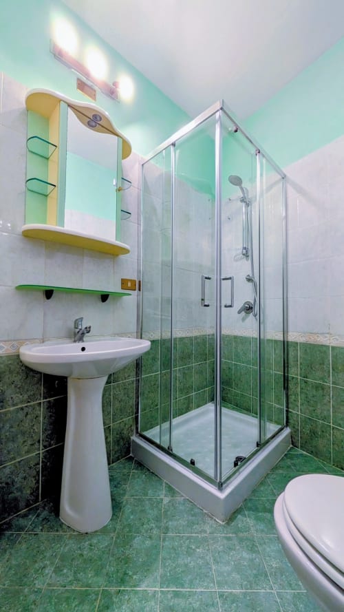 Modern bathroom with green tile, pedestal sink, glass shower enclosure, toilet, and yellow shelving in Villa Italia apartment