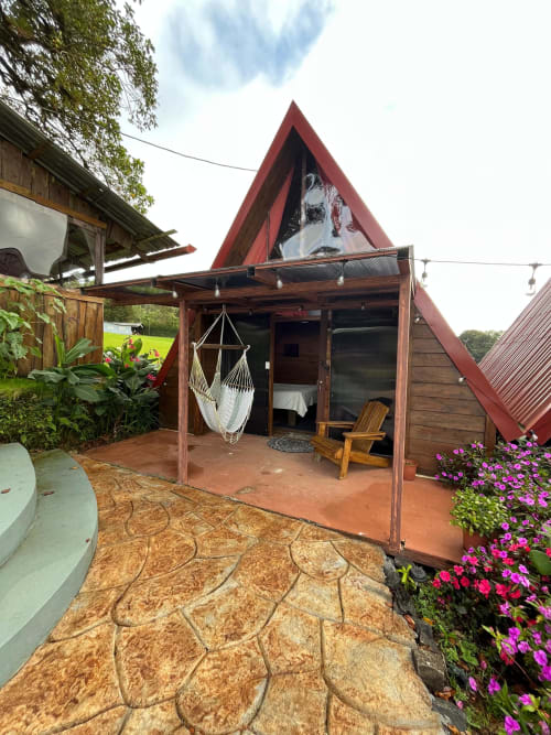 Red triangular chalet with wooden porch, hammock, and pink flowers at Chalets Vistas del Poas in Heredia, Costa Rica