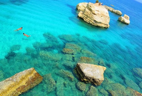 Vista aerea delle acque turchesi cristalline del Mediterraneo con snorkelisti, affioramenti rocciosi e formazioni subacquee