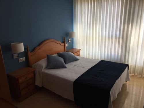 Dormitorio doble en Pensión San Vicente con cama de madera, ropa de cama gris, paredes azules, lámpara y vistas a la montaña a través de las cortinas
