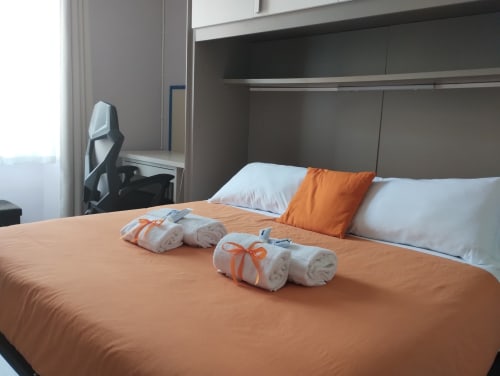 Camera da letto moderna con dettagli arancioni, asciugamani arrotolati, cuscini bianchi, area scrivania e scaffalature presso Co-Host Now a Roma