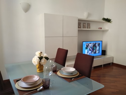 Zona pranzo di moderno appartamento a Roma con tavolo preparato, sedie bordeaux, TV e eleganti mobili bianchi