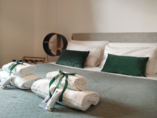 Camera da letto elegantemente arredata con asciugamani arrotolati legati con nastro verde, testata grigia, cuscini bianchi e puf verde scuro