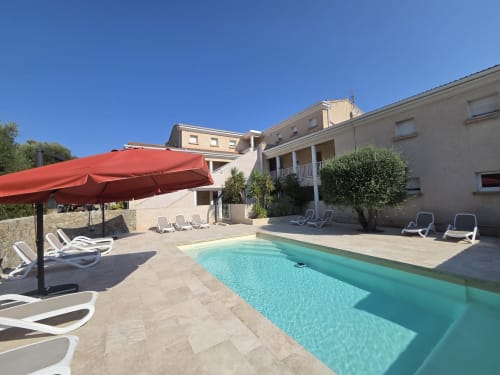 Piscine de la Résidence Saint Michel aux eaux turquoise, parasol rouge, transats et bâtiments en pierre beige à L'île Rousse