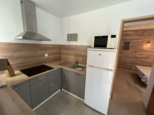 Modern kitchen at Le Maquis Plage hotel with gray cabinetry, wooden backsplash, white appliances, and open door to bedroom