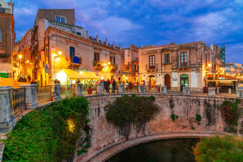 Piazza storica siciliana al tramonto con edifici barocchi illuminati, ponte di pietra e muri ricoperti di edera vicino a Fontane Bianche