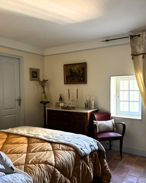 Elegant bedroom at Domaine Du Commandeur with gold quilted bed, dark dresser, period artwork, and garden views in Mondragon
