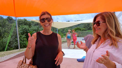 Two women smiling under orange shade at Corse et Zen, with guests and green hills visible in Taglio Isolaccio