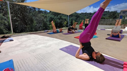 Yoga class on outdoor terrace at Corse et Zen with instructor in headstand, purple mats, shade sail, and forested Corsican