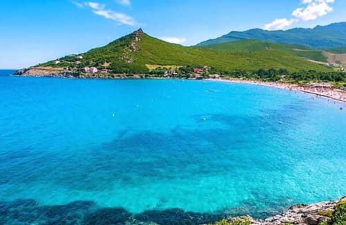 Turquoise Mediterranean beach with green mountains and coastal village in Corsica