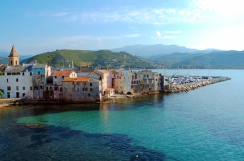 Colorful waterfront buildings in Corsica with turquoise Mediterranean water, mountains, and church bell tower at Taglio
