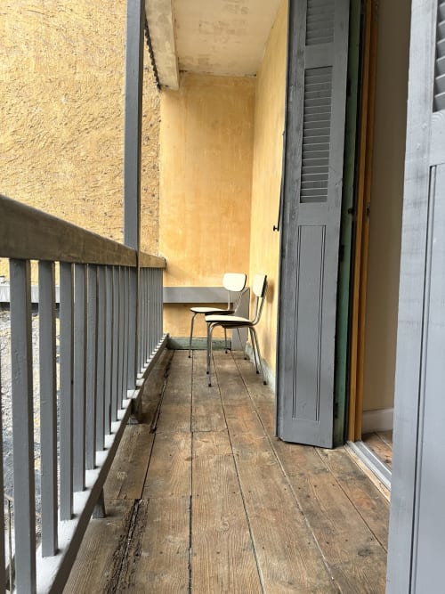 Wąski balkon w Ombre de la Tour w Lourdes z drewnianym tarasem, metalową barierką i dwoma krzesłami z widokiem na żółtą kamienną ścianę