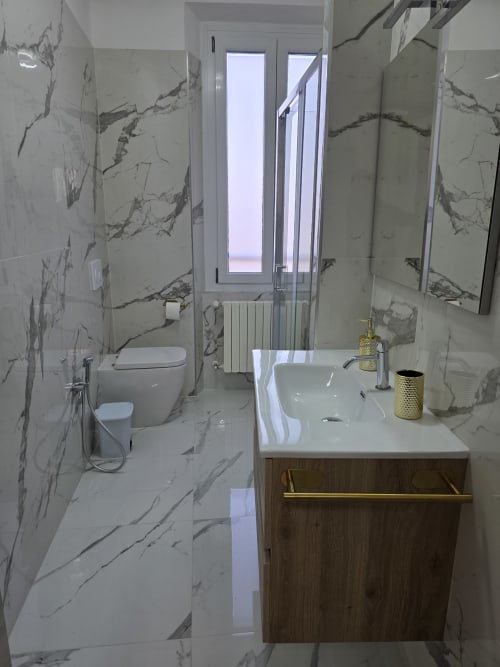 Bagno moderno con rivestimenti effetto marmo, lavabo bianco con base in legno, WC e doccia in vetro presso THE V