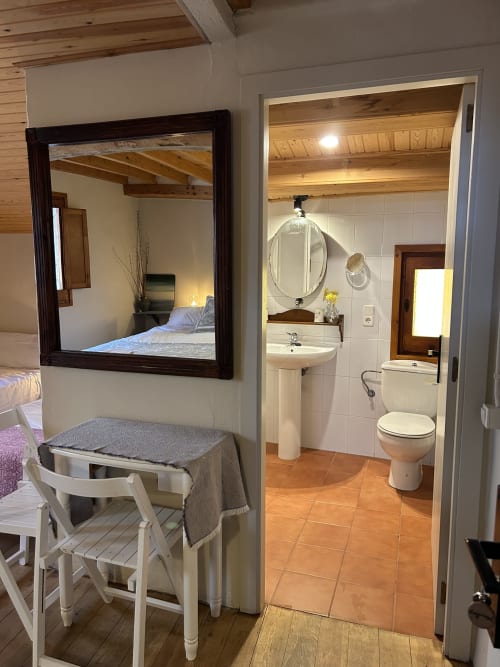 Habitación L'Esquirol en Casa Tapiolas con cama, espejo, silla blanca y baño privado con azulejos de terracota