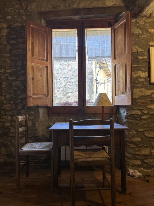 Habitación rústica de piedra en Casa Tapiolas con contraventanas de madera, mesa, sillas y vistas a las montañas en Molló, Girona