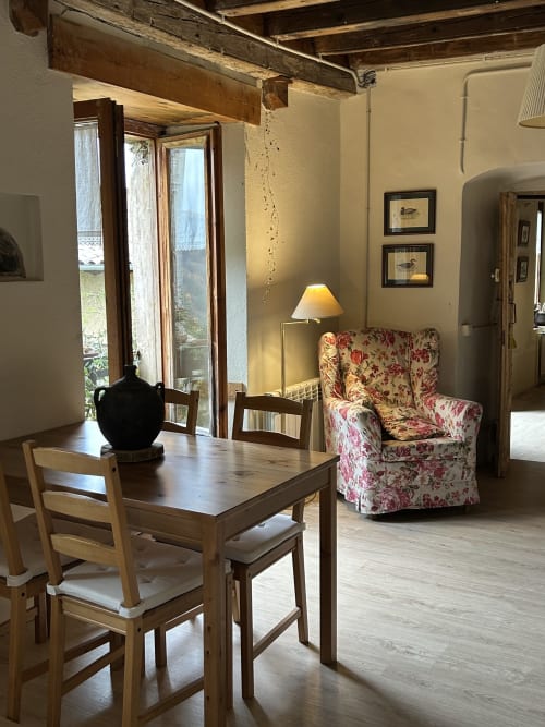 Acogedor rincón de estar en Casa Tapiolas en Molló con sillón floral, mesa de comedor de madera y puertas con vistas a Girona