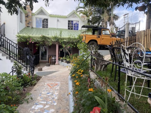 Cour de la maison d'hôtes écologique Baz-Art à Rodrigues avec bâtiment blanc, toiture verte, camion orange, plantes fleuries et espaces de détente
