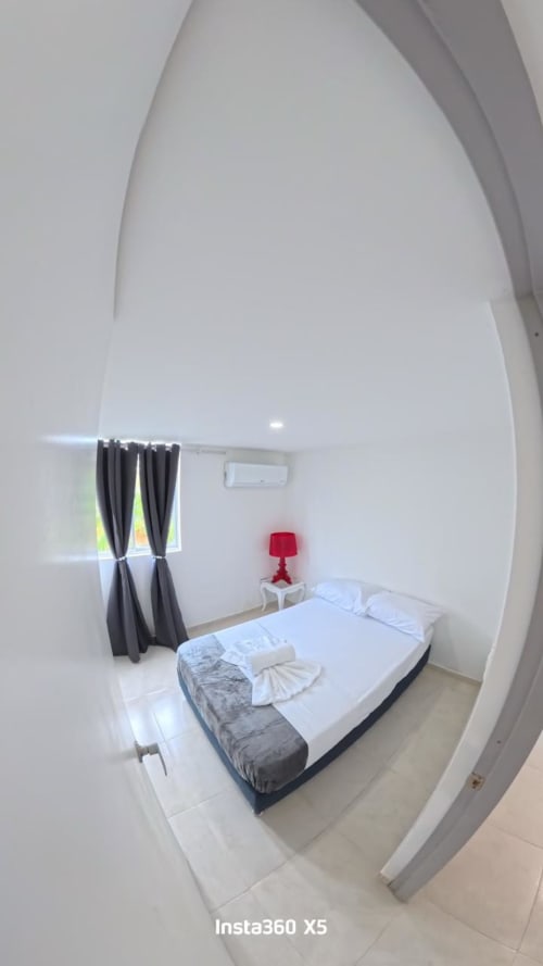 Moderno dormitorio en Olamar Living de Santiago de Tolú con cama queen, lámpara roja, cortinas oscuras y paredes blancas
