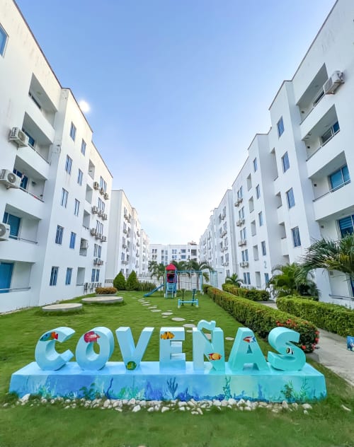 Complejo de apartamentos Coveñas Retreat de Olamar Living con edificios blancos, jardín verde y letrero colorido "COVEÑAS"