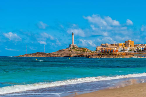 Vista de la playa costera de Blanca, España con aguas turquesas, arena blanca, faro en península rocosa y colorido