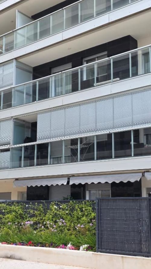 Edificio moderno en Benicàssim con balcones acristalados, vistas al jardín, plantas floridas y toldo negro