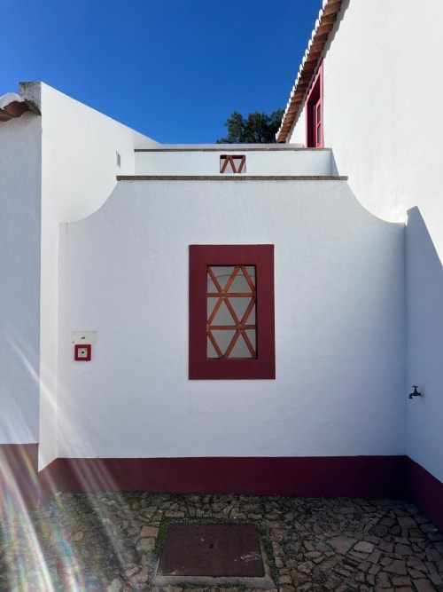 Entrada de villa portuguesa com paredes brancas, porta com moldura de madeira vermelho profundo e janela com treliça geométrica em Casas de Juromenha,