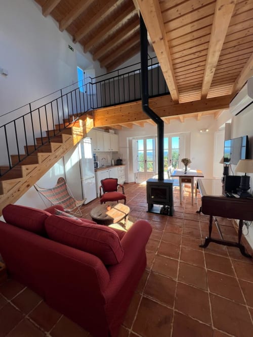 Área de estar em plano aberto na villa Casa dos Queixos, Marvão com vigas de madeira, sofá vermelho, lareira, escadas e vistas para a montanha