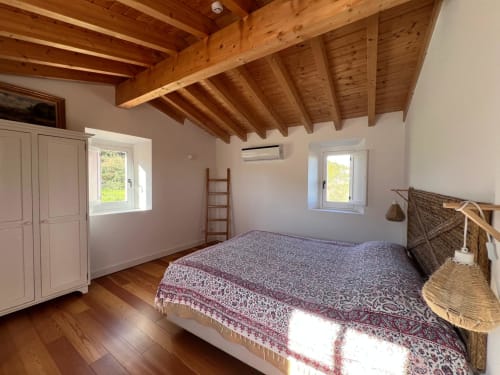 Quarto espaçoso com vigas de madeira aparente, cama queen, guarda-roupa rosa e janelas com vista para o jardim na Casa dos Queixos