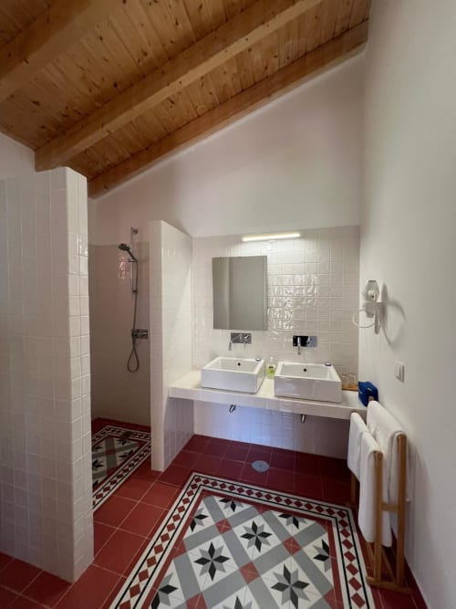 Banheiro moderno em Casas de Juromenha com dois lavatórios, chuveiro aberto, piso com azulejos decorativos e vigas de madeira aparente