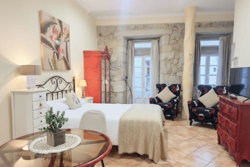 Dormitorio del apartamento Vermello en Baiona con cama, muros de piedra, sillones, mobiliario rojo y balcón con vistas a la ciudad