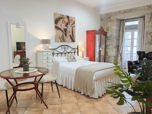 Elegante dormitorio en Holibai Baiona con cama de hierro forjado, ropa blanca, mesa redonda y paredes de piedra con balcón