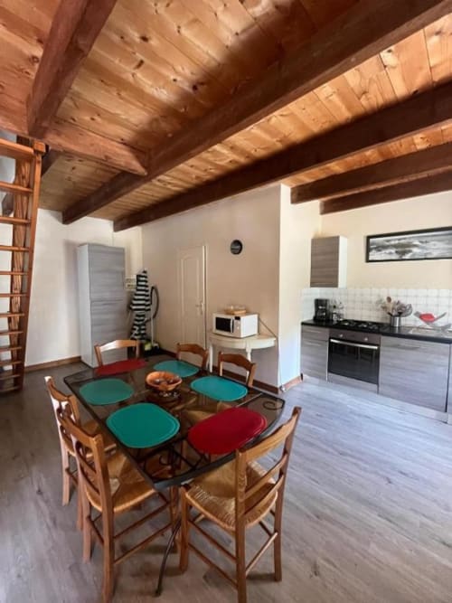 Salle à manger avec poutres en bois, table entourée de chaises bleu-vert et rouge, et cuisine dans la villa bretonne Le Gîte