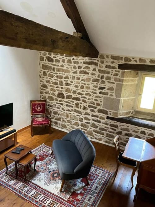 Salon Suite Seigneuriale aux murs de pierre, poutres apparentes, fauteuil moderne noir et mobilier en bois au Manoir de