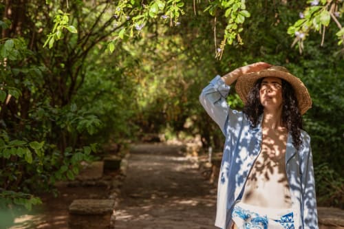 Woman in straw hat and linen shirt walks through lush tree-lined path at Case Zucco B&B in Terrasini, Italy