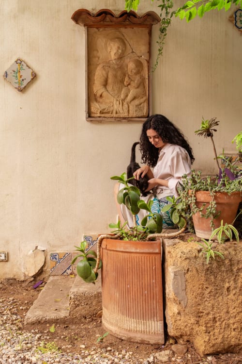 Woman tending to potted plants in courtyard at Case Zucco B&B in Terrasini, Italy, with terracotta pots and traditional wall