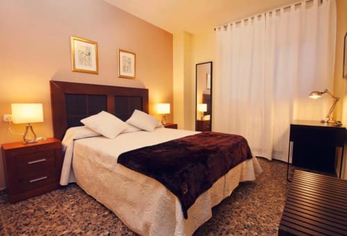 Elegante dormitorio en Gestyp-AT en Mérida con cabecero burdeos, ropa de cama blanca, iluminación cálida y cortinas con vista a la ciudad