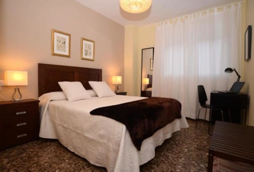 Dormitorio moderno en apartamento de Mérida con cama de madera oscura, ropa blanca, manta marrón, mesitas con lámparas y escritorio