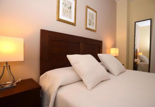 Acogedora habitación en apartamento Gestyp-AT en Mérida con cabecera de madera oscura, almohadas blancas, iluminación cálida y decoración botánica