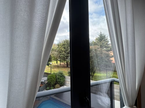 Vista desde el balcón del apartamento Cousiña en Baiona con piscina y jardines exuberantes con árboles, cortinas blancas enmarcan la ventana