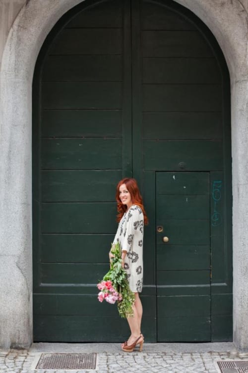 Donna con capelli rossi che tiene fiori rosa davanti all'ingresso di Alessia's Place B&B a Como, Italia, di fronte a grandi porte ad arco verdi