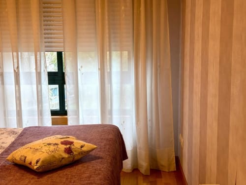 Dormitorio luminoso con almohada amarilla en la cama, cortinas semitransparentes y vistas a la ciudad en el apartamento Angelitiña de Baiona
